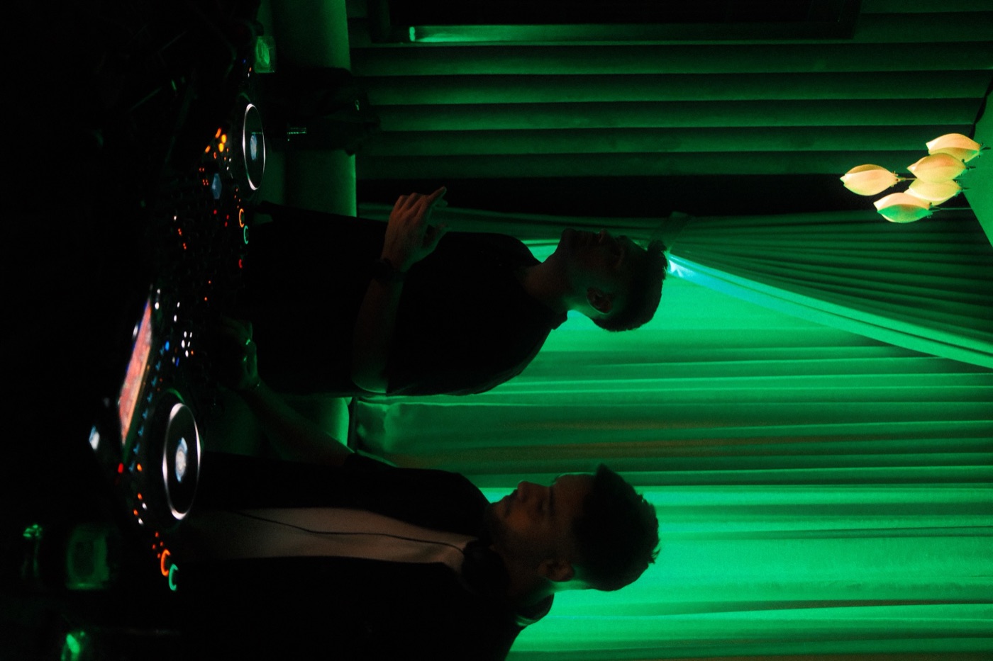 Two DJ silhouettes against green-lit curtains